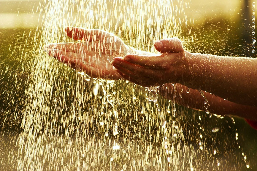 Ein paar Hände sind mit der Handfläche nach oben gedreht. Ein Wasserstrahl trifft auf die Hände. 